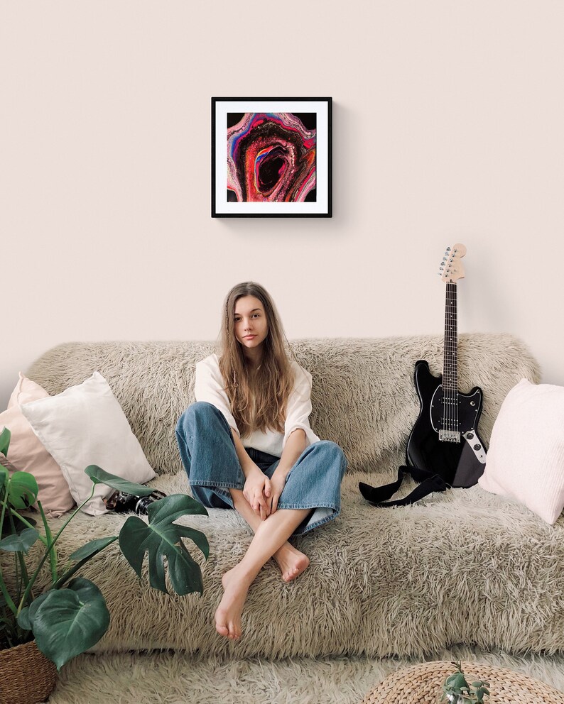 May include: A young woman sits on a fluffy, light brown couch with a black electric guitar leaning against the couch. She is wearing a white long-sleeved shirt and blue jeans. A framed abstract painting with pink, red, blue, and black colors hangs on the wall behind her.