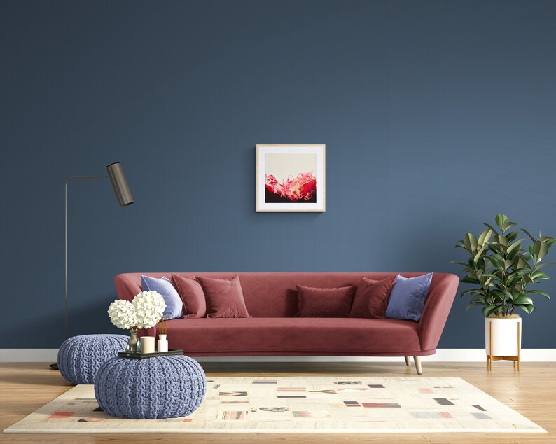 May include: A red velvet sofa with blue cushions sits in a living room with a blue wall, a white rug, a blue knitted footstool, and a potted plant. A framed abstract painting hangs on the wall above the sofa.