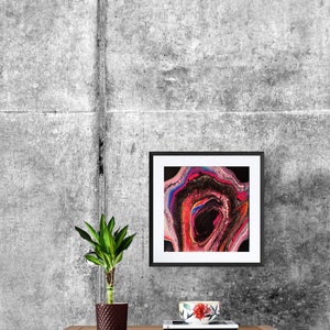 May include: A framed abstract painting with swirling colors of pink, blue, red, and black. The painting is hanging on a gray concrete wall above a wooden table with a potted plant and a cup.