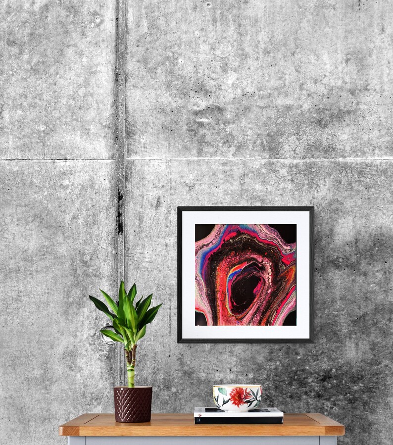 May include: A framed abstract painting with swirling colors of pink, blue, red, and black. The painting is hanging on a gray concrete wall above a wooden table with a potted plant and a cup.