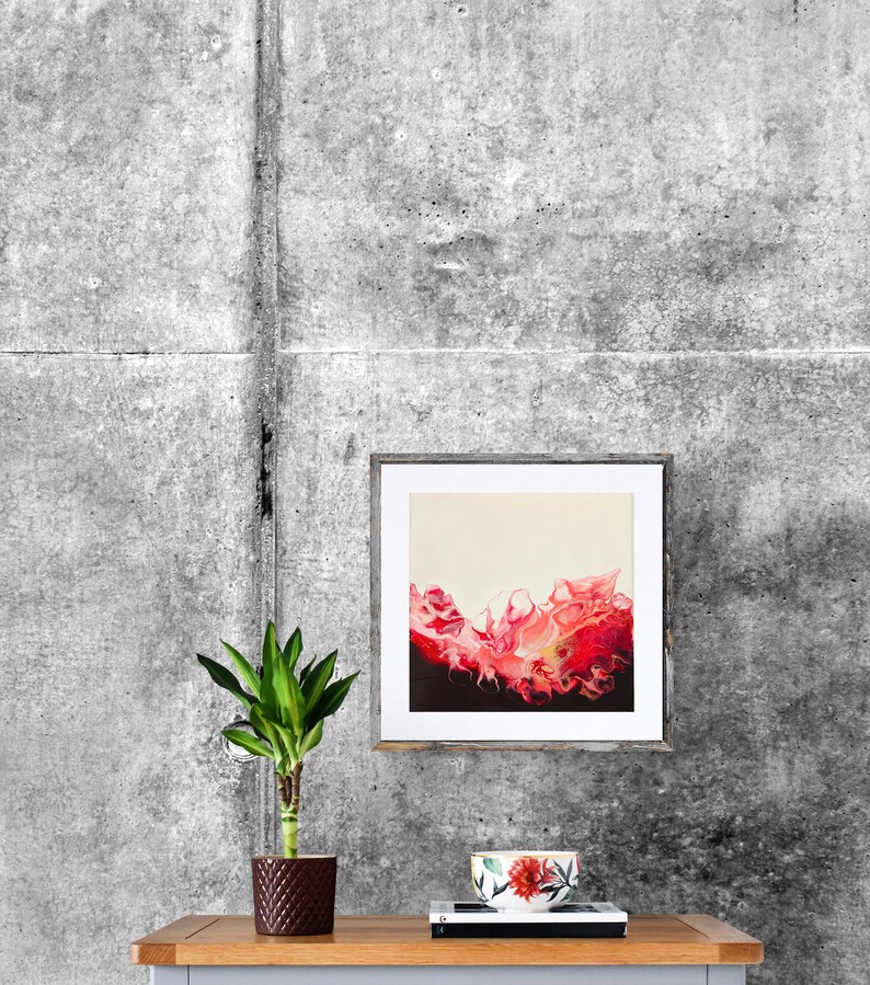May include: A framed abstract painting with a pink and red wave-like design on a cream background. The painting is hanging on a grey concrete wall above a wooden table with a potted plant and a floral patterned cup.