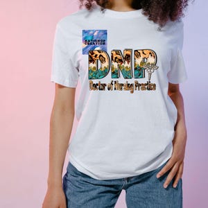Può includere: Maglietta bianca con una grafica colorata che dice "DNP Doctor of Nursing Practice" con un simbolo di caduceo.