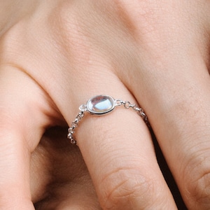 May include: A silver chain ring with a clear oval-shaped gemstone. The ring is on a person's finger.