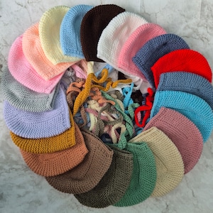 May include: A collection of 20 crocheted baby bonnets in various colours, including pink, blue, red, brown, and yellow. The bonnets are arranged in a circle, with the ties of each bonnet visible.