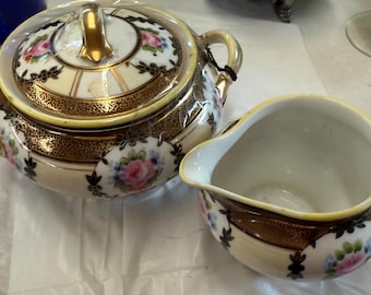 May include: A vintage porcelain sugar bowl with a lid and a creamer. The set features a cream-colored base with gold trim and floral accents in pink, blue, and white. The sugar bowl has a gold handle and a matching lid.