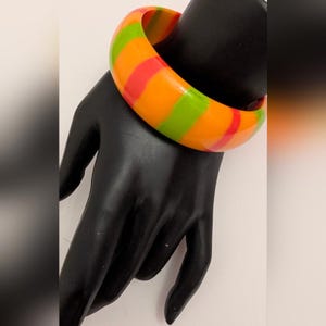 Peut inclure: Un bracelet jonc coloré avec un motif rayé orange, vert et rouge. Le bracelet large et épais est présenté sur un support à bijoux en forme de main noire. Un accessoire de mode audacieux.