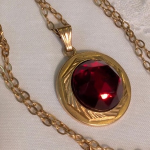 May include: A gold-toned necklace featuring a round pendant with a faceted deep red stone at its centre. The pendant has a textured, geometric border and is suspended from a matching gold-toned chain with oval-shaped links.
