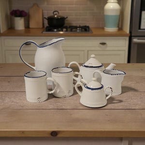 May include: A white enamelware tea set with blue edging. The set comprises a jug, teapot, sugar bowl, creamer, and two mugs. The set is displayed on a wooden table, with a kitchen in the background.