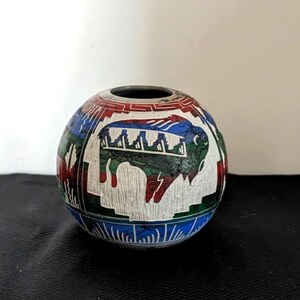 May include: A round, hand-painted ceramic vase with a black opening. The vase features a white background with a detailed design of a bison, geometric shapes, and Native American-inspired patterns in red, blue, and green.