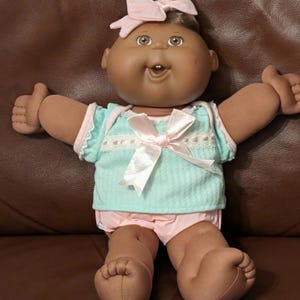 May include: A vintage Cabbage Patch Kid doll with dark skin, brown hair, and brown eyes. The doll is wearing a light blue top with pink trim and a pink bow, and pink shorts. The doll's arms are outstretched.