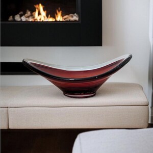 May include: A decorative, red glass bowl with a wide, curved design. The bowl has a clear rim and sits on a beige upholstered bench. A fireplace with a black frame and a burning fire is in the background.
