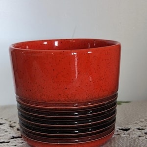 May include: A ceramic plant pot with a glossy red upper section and a lower section featuring horizontal bands of dark brown and black. The pot has a speckled texture and a smooth, rounded rim.