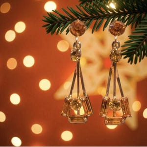 May include: Gold-coloured earrings with amber gemstones. The earrings hang from a green pine branch against a warm, blurred background with bokeh lights. The earrings have a geometric design with multiple facets.