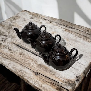 Peut inclure: Trois théières noires brillantes avec des corps arrondis, des becs verseurs et des anses en boucle sont disposées sur une surface en bois vieilli. Chaque théière a un couvercle assorti avec un petit bouton. Les théières sont de tailles variables.