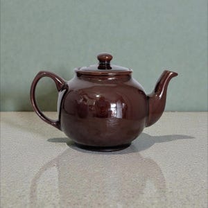 May include: A brown ceramic teapot with a lid, handle, and spout. The teapot has a glossy finish and is on a light-coloured surface. The teapot is designed for brewing tea.