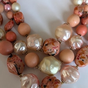 May include: A multi-strand necklace featuring a variety of beads in shades of peach, cream, and coral. The beads include smooth round spheres, textured pearlescent shapes, and irregular speckled pieces, all connected by small gold-tone metal caps.