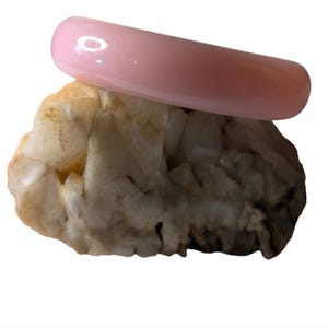 May include: A pale pink, curved bangle bracelet sits on a rough, beige and white rock. The bracelet has a smooth, glossy finish. The rock has a natural, textured appearance with variations in colour and surface.