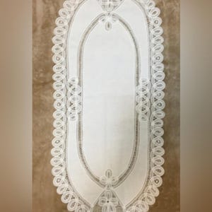 May include: White oval-shaped lace table runner. The runner features a detailed floral and openwork design along the edges and center. The lace is intricately patterned, creating a delicate and elegant aesthetic. The runner is placed on a brown surface.