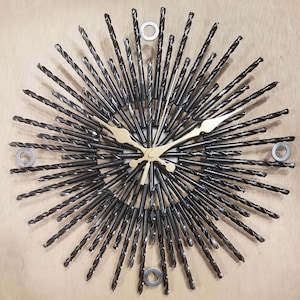 May include: A unique wall clock with a sunburst design made from black drill bits. The clock features gold-colored hands and a central clock face. The clock is mounted on a light-colored wooden surface, with three silver washers.