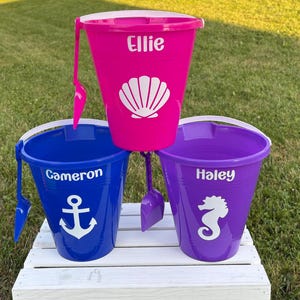 May include: Three colorful beach buckets with matching shovels. The pink bucket has a seashell graphic and the name "Ellie". The blue bucket has an anchor graphic and the name "Cameron". The purple bucket has a seahorse graphic and the name "Haley".