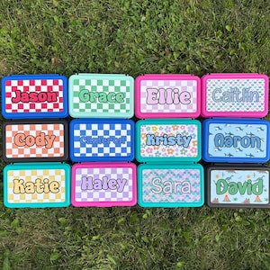 May include: A collection of colorful personalized lunch boxes with names like Jason, Grace, and Ellie. Each box features a unique pattern and color scheme, including checkered and floral designs. The boxes are arranged on a bed of green grass.