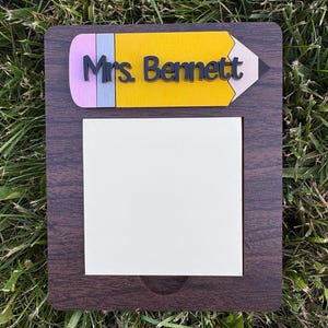 May include: A wooden teacher's nameplate with a pencil design. The top of the nameplate is shaped like a yellow pencil with a pink eraser and a gray pencil lead. The name "Mrs. Bennett" is in black letters. A white notepad is below.