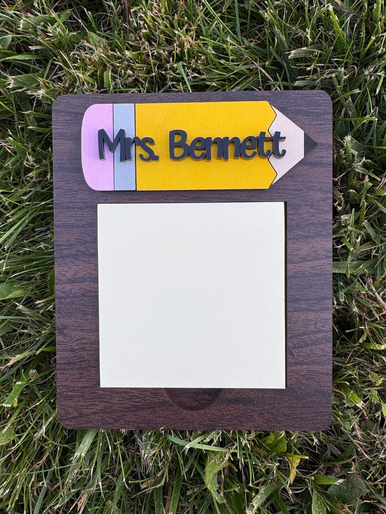 May include: A wooden teacher's nameplate with a pencil design. The top of the nameplate is shaped like a yellow pencil with a pink eraser and a gray pencil lead. The name "Mrs. Bennett" is in black letters. A white notepad is below.