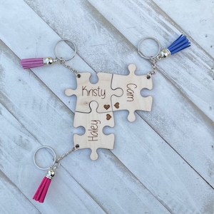 May include: Three wooden puzzle pieces with keychains and tassels. The pieces are connected and have the names "Kristy", "Cam", and "Haley" engraved on them. The tassels are pink and blue.