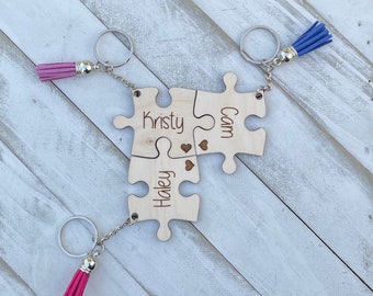 May include: Three wooden puzzle pieces with keychains and tassels. The pieces are connected and have the names "Kristy", "Cam", and "Haley" engraved on them. The tassels are pink and blue.