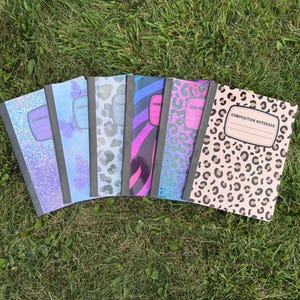 May include: Six composition notebooks arranged in a fan shape, featuring various patterns including holographic glitter, purple butterflies, grey leopard print, pink and blue zebra stripes, blue leopard print, and a tan leopard print with a name label.