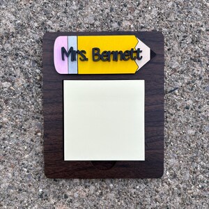 May include: A dark brown wooden notepad holder with a yellow pencil design at the top. The pencil has a pink eraser, a yellow body, and the name "Mrs. Bennett" in black. A square pad of sticky notes is attached.