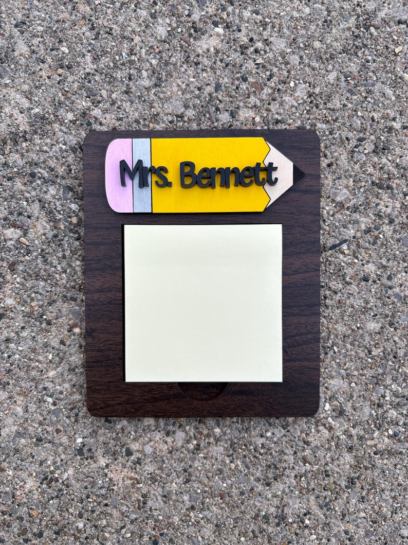 May include: A dark brown wooden notepad holder with a yellow pencil design at the top. The pencil has a pink eraser, a yellow body, and the name "Mrs. Bennett" in black. A square pad of sticky notes is attached.