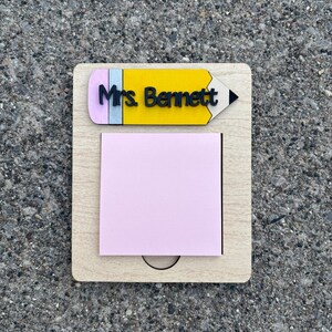 May include: Wooden desk organizer with a pencil design and the name "Mrs. Bennett". The organizer has a yellow pencil top with a black tip and a pink eraser. A pink sticky note pad is attached to the wooden base.