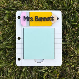 May include: A white notepad with a light blue sticky note and a yellow pencil-shaped topper. The pencil has a pink eraser and the name "Mrs. Bennett" printed on it. The notepad has a spiral notebook design.