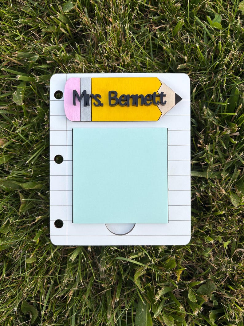 May include: A white notepad with a light blue sticky note and a yellow pencil-shaped topper. The pencil has a pink eraser and the name "Mrs. Bennett" printed on it. The notepad has a spiral notebook design.