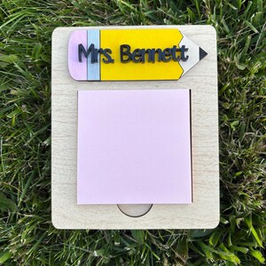 May include: A wooden desk organizer with a light wood finish. It features a yellow pencil-shaped nameplate with the text "Mrs. Bennett" in black. A pink notepad is attached to the organizer.