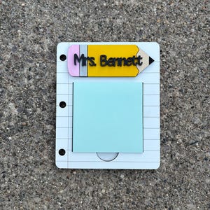 May include: A white notepad-shaped wooden desk organizer with a yellow pencil topper that reads "Mrs. Bennett." The notepad has a light blue sticky note and three black binder holes.
