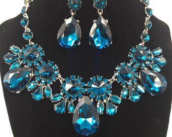 May include: A dazzling jewelry set featuring a necklace and matching earrings. The necklace and earrings are adorned with large, faceted, teal-colored gemstones set in a silver-toned metal. The necklace has a bib design with a large central teardrop stone.