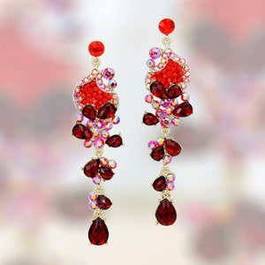 May include: A pair of long, dangling earrings with red and pink crystals. The earrings are made of gold-toned metal and feature a cluster of red and pink crystals at the top, followed by a series of smaller crystals that dangle down.