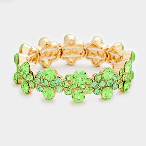 May include: A gold stretch bracelet with green crystal embellishments. The bracelet is made of small, round crystals that are clustered together in a flower-like pattern.
