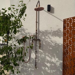 May include: An outdoor shower system made of copper-toned metal pipes, featuring a circular shower head at the top, a vertical pipe assembly, two manual control valves, and a flexible handheld shower hose attachment mounted on a wall.