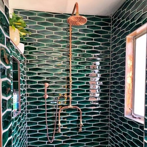 May include: A contemporary shower room with dark green, elongated hexagonal tiles. The shower has copper fixtures and a showerhead. A small window with a wooden frame is visible, along with a small plant in a pot.