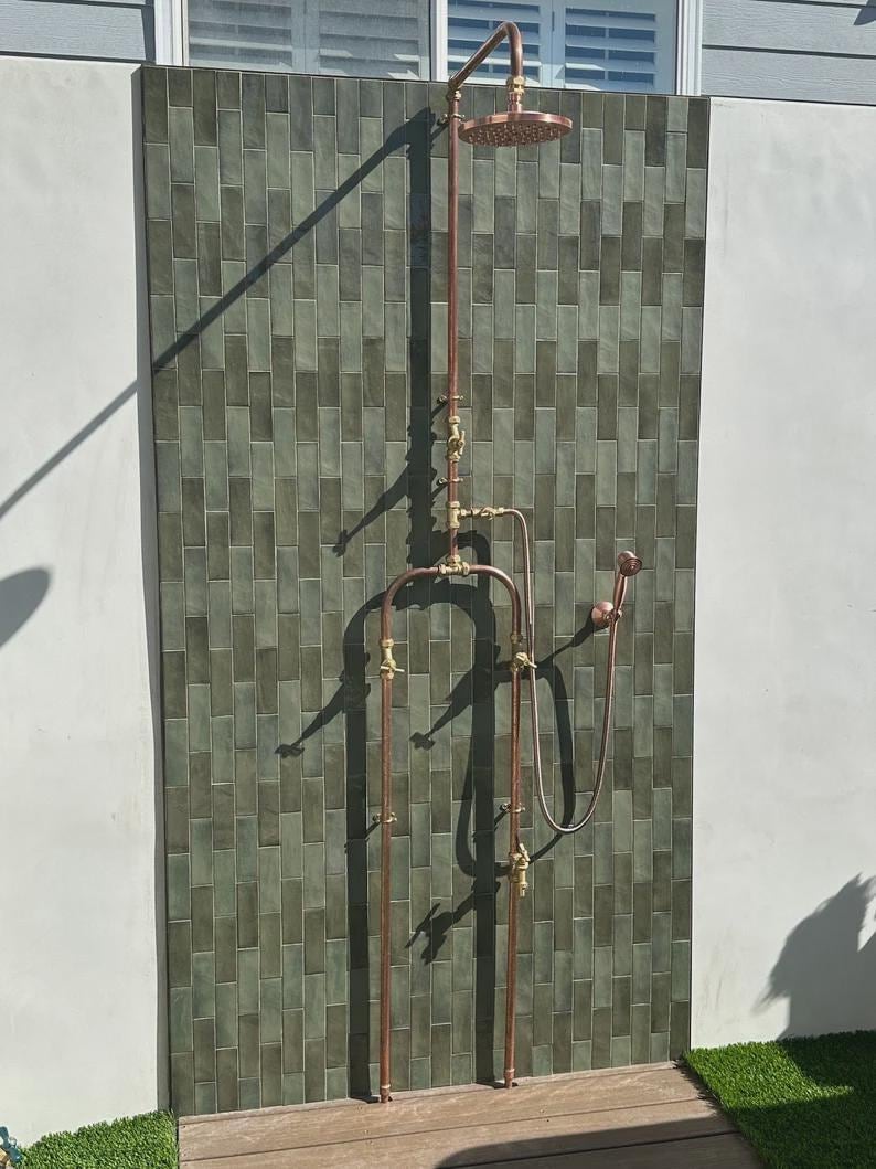 May include: Outdoor shower fixture with copper pipes and fittings, including a rain shower head and handheld shower. The shower is mounted on a green tiled wall. The fixture has a vintage, industrial design.