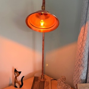 May include: A table lamp featuring a copper pipe stem and a wide, bowl-shaped copper shade. The lamp is mounted on a rectangular wooden base composed of alternating light and dark wood strips. A single warm-toned light bulb is visible inside.