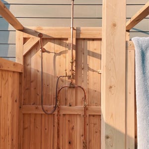 May include: Outdoor shower with copper shower head and piping, set against a wooden structure. The shower head is round with multiple holes. The wooden door and light blue wall provide a natural backdrop. The shower is a rustic design.