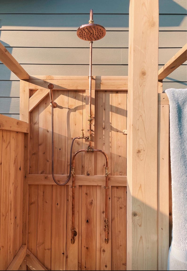 May include: Outdoor shower with copper shower head and piping, set against a wooden structure. The shower head is round with multiple holes. The wooden door and light blue wall provide a natural backdrop. The shower is a rustic design.