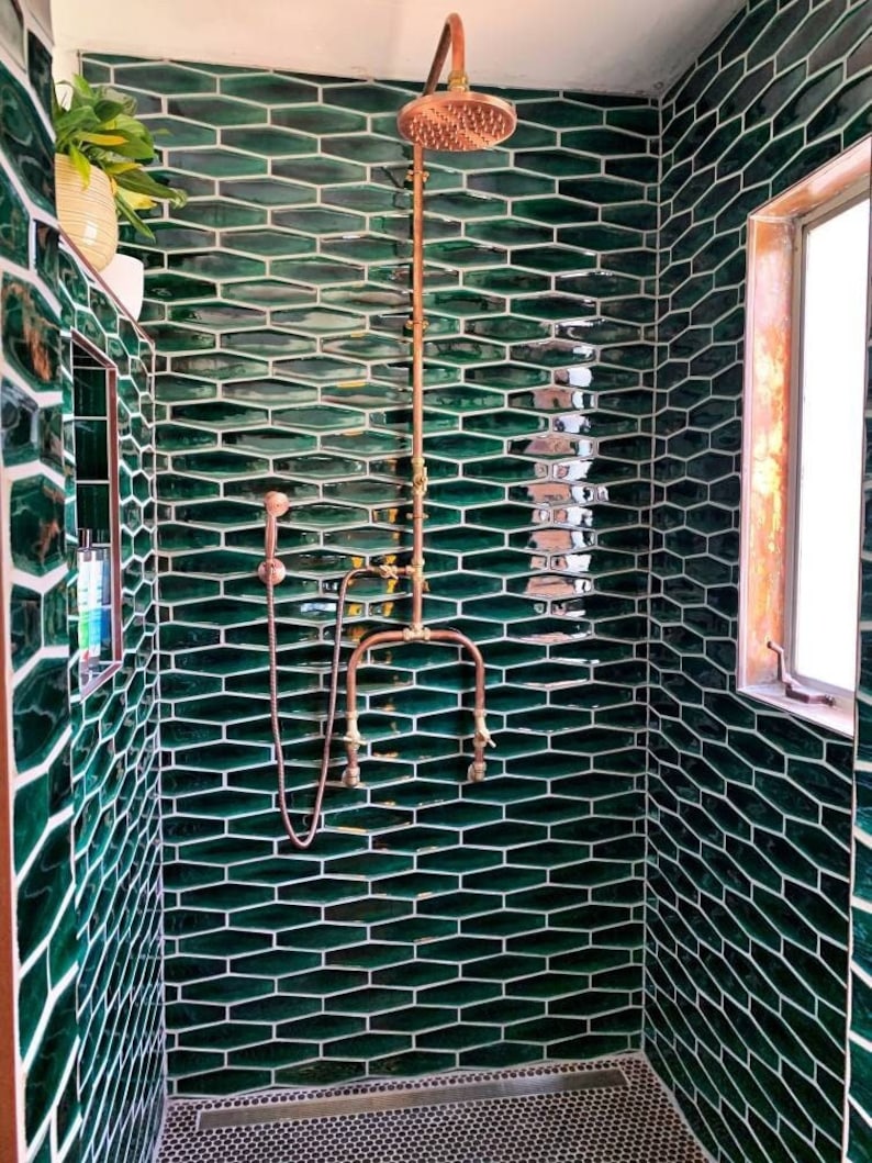 May include: A contemporary shower room with dark green, elongated hexagonal tiles. The shower has copper fixtures and a showerhead. A small window with a wooden frame is visible, along with a small plant in a pot.