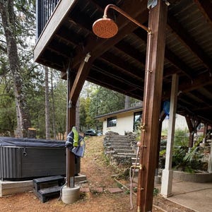 May include: An outdoor shower featuring a copper showerhead and fittings, attached to a wooden post. A hot tub is visible on the left, with a house in the background. The shower is beneath a wooden deck.