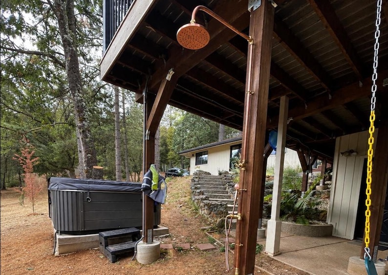 May include: An outdoor shower featuring a copper showerhead and fittings, attached to a wooden post. A hot tub is visible on the left, with a house in the background. The shower is beneath a wooden deck.