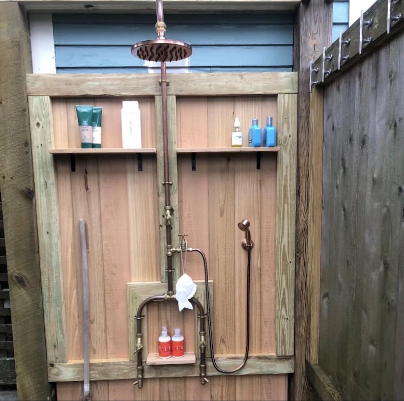 May include: Outdoor shower featuring copper piping and a rain shower head. The shower is integrated into a wooden structure with shelves holding toiletries. A handheld shower head is also included. The shower is enclosed by wooden walls.